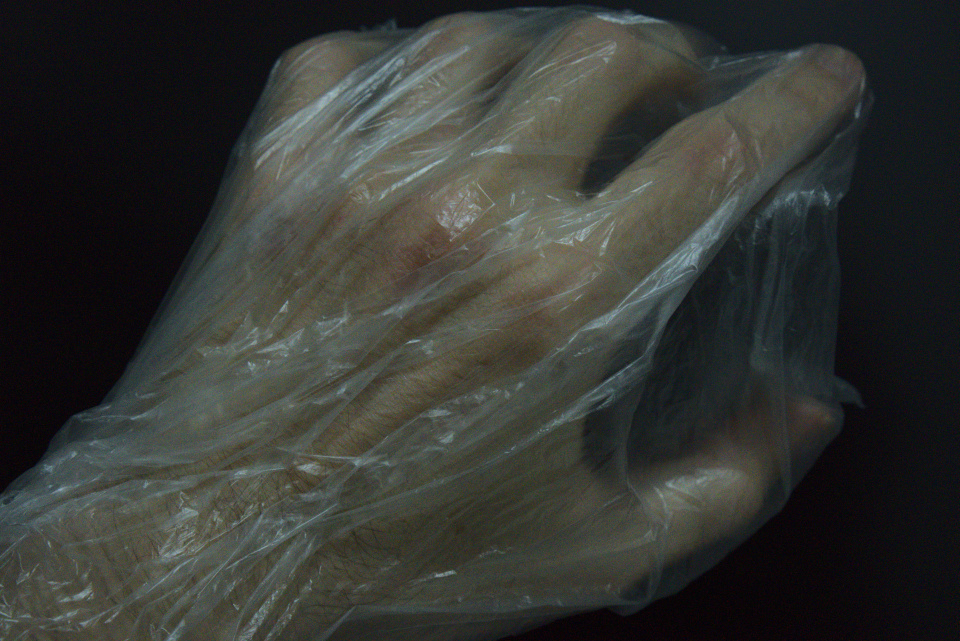 Uma mão embalada em uma sacola plástica transparente em vácuo. A mão, de aparência mórbida, estendida procurando algo.