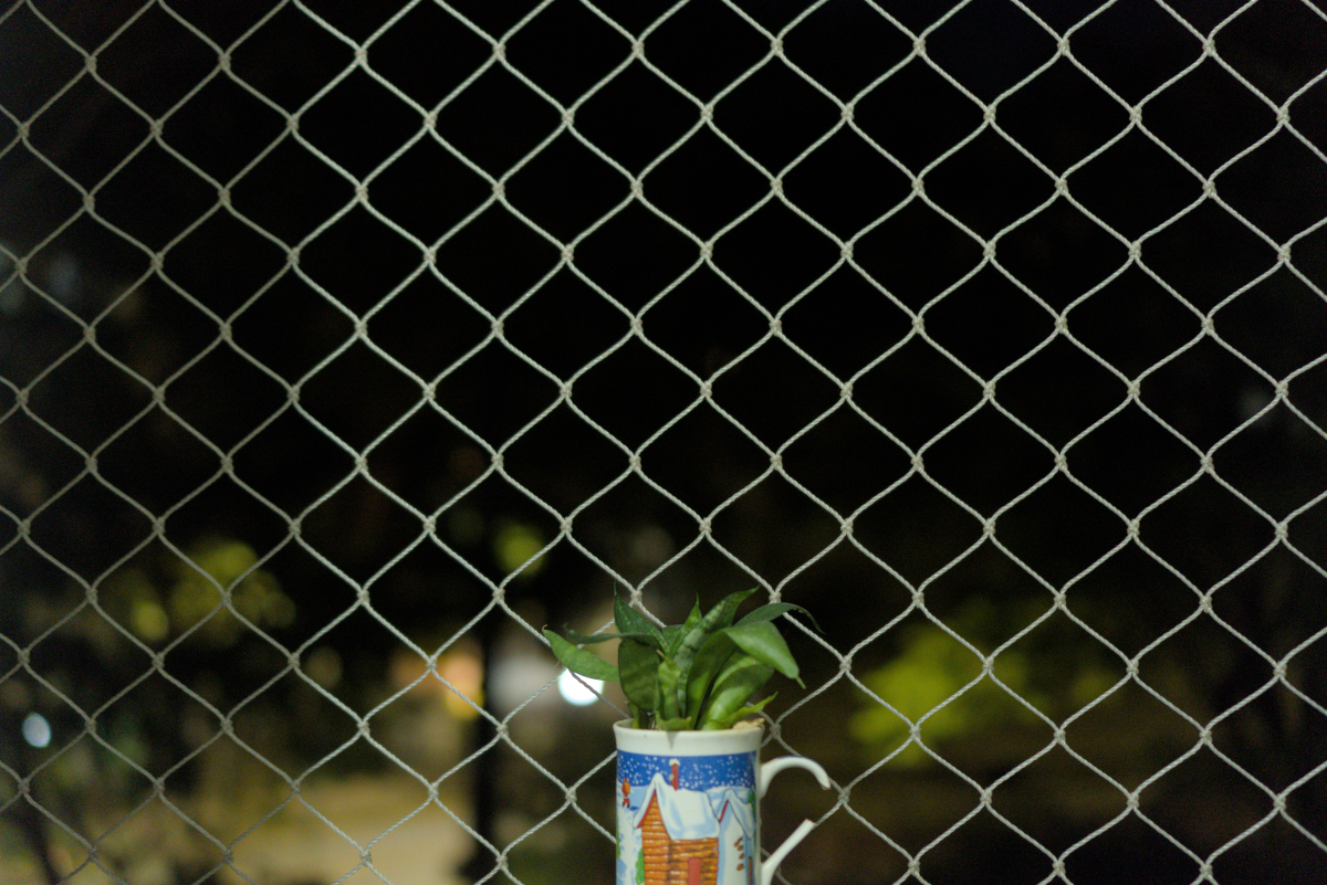 Ao centro, na parte de baixo, uma caneca com a asa quebrada e uma ilustração natalina. Atrás uma rede de proteção, possivelmente de uma janela. Ao fundo, borrado, as luzes noturnas da cidade.