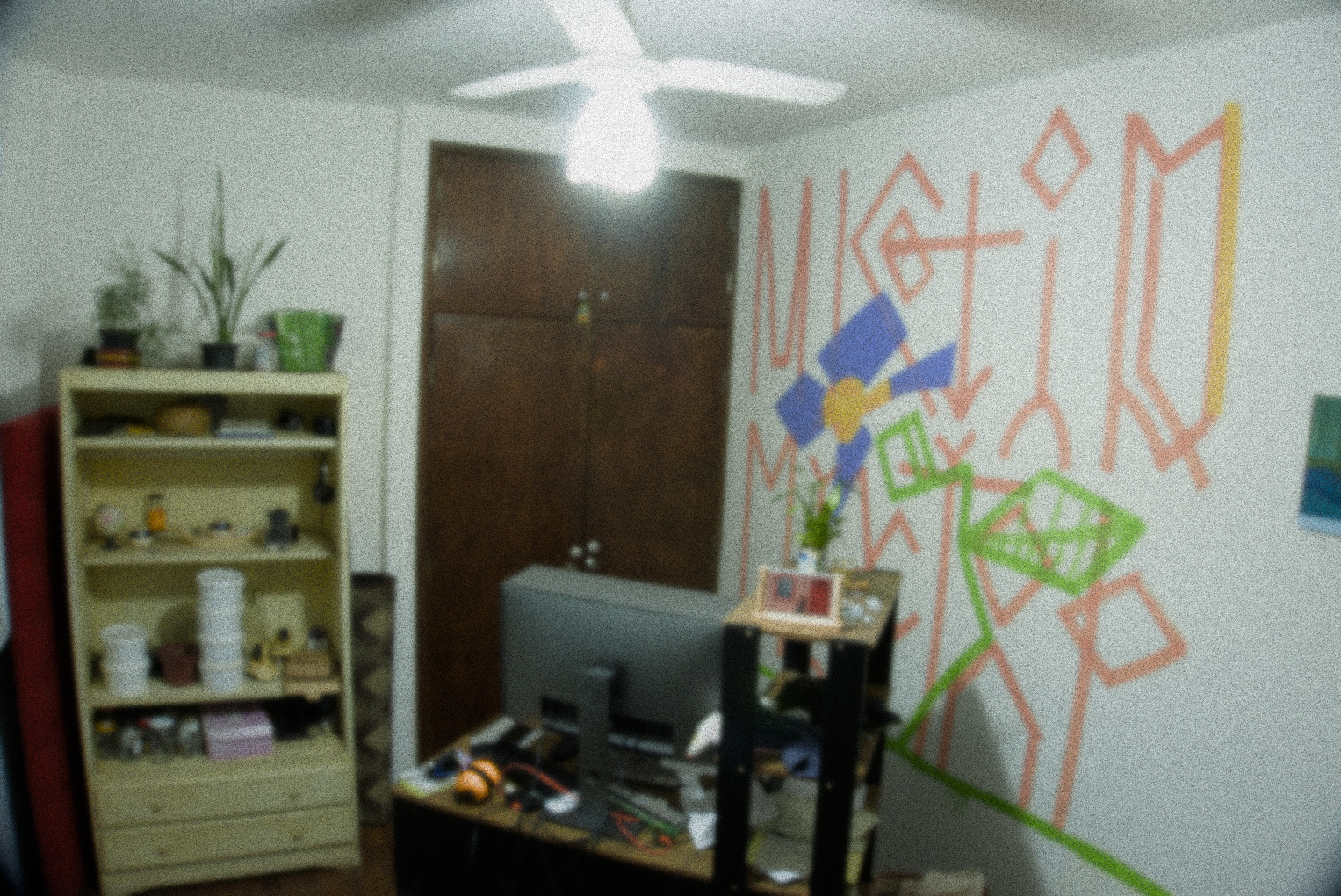 Uma mesa de computador vista por trás. Muitas coisas jogadas sobre a mesa, mas a imagem não possui nitidez para identificar os objetos. Além da mesa uma pixação na parede.